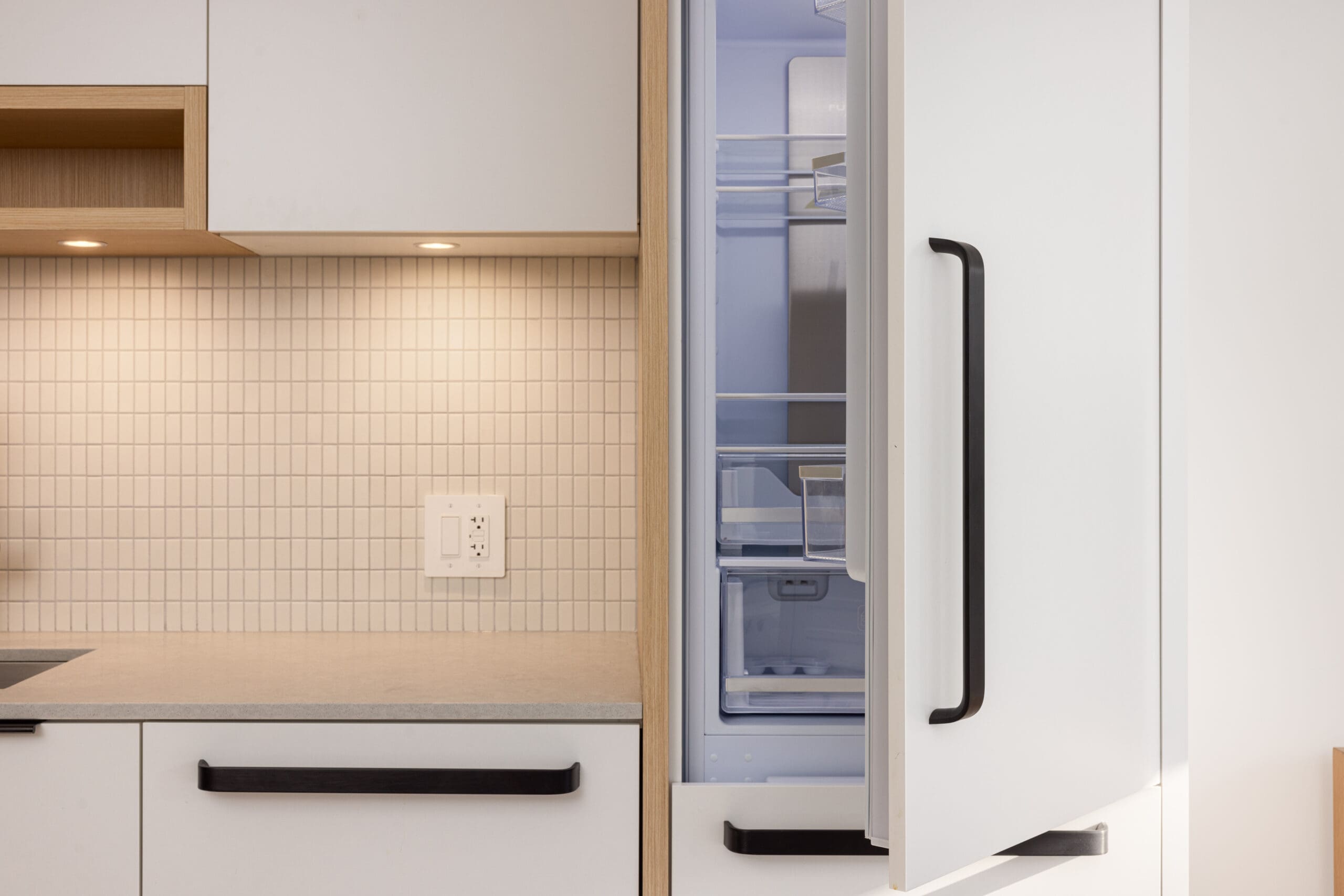 Open refrigerator door in a modern kitchen, showing empty shelves and drawers. Cream-tiled backsplash with white cabinets and black handles. Under-cabinet lighting is turned on.