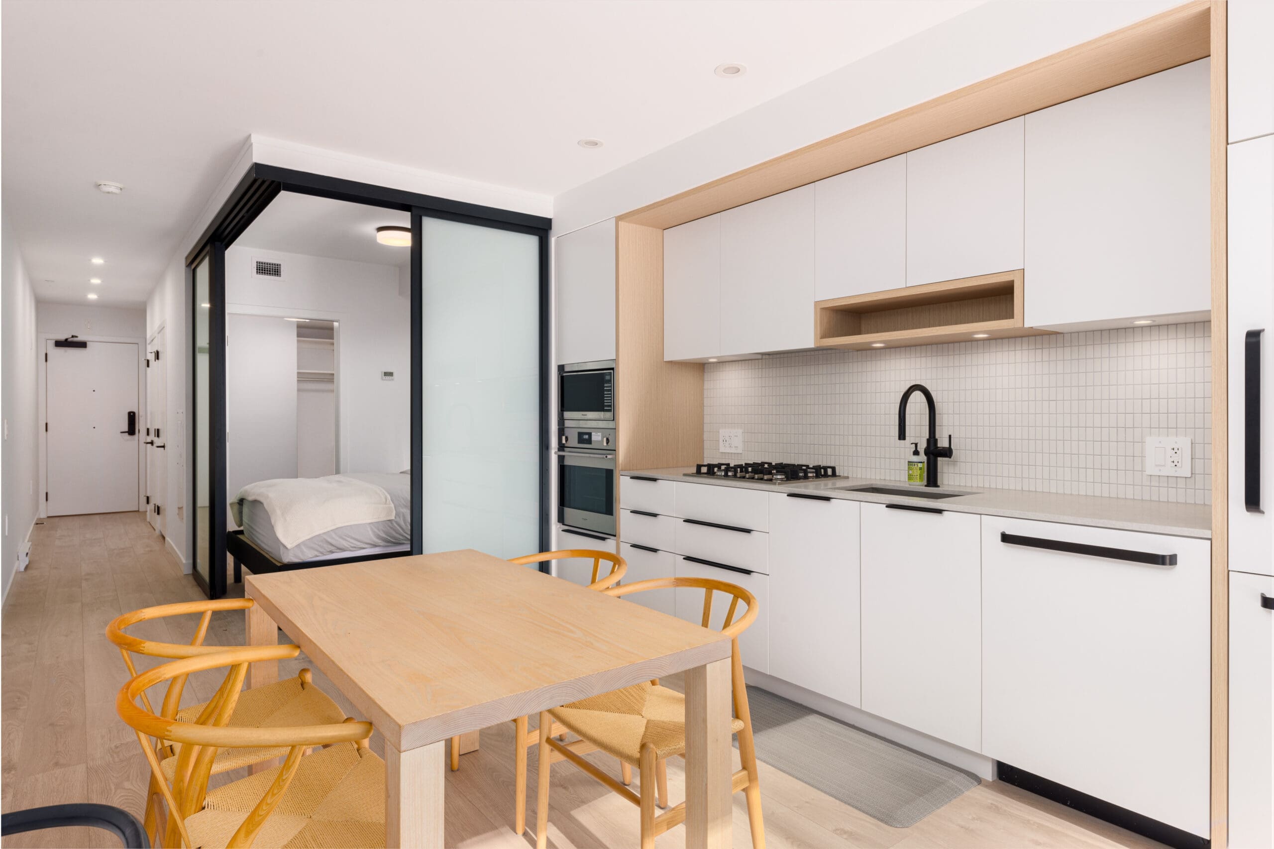 Modern kitchen with white cabinetry, built-in appliances, and a black faucet. A wooden dining table with four chairs sits in the foreground. A bedroom is visible through sliding glass doors.