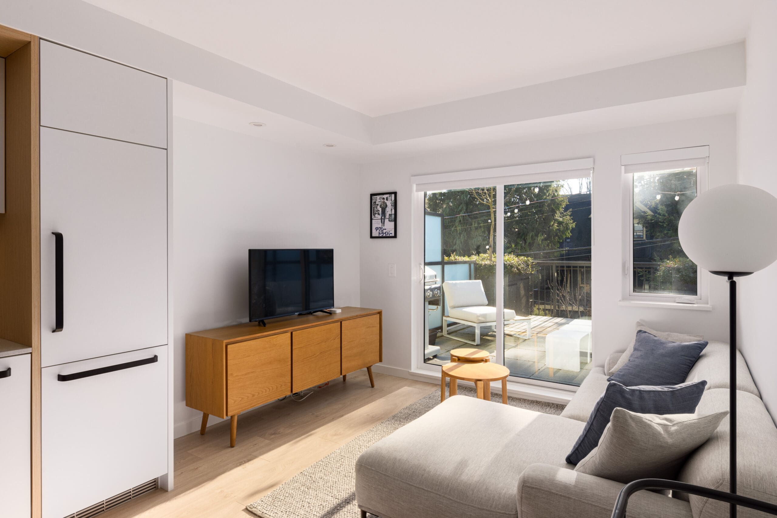 Modern living room with a TV on a wooden stand, a gray sectional sofa, and a floor lamp. A glass door leads to a balcony with outdoor seating. Light-colored walls and wooden floor.