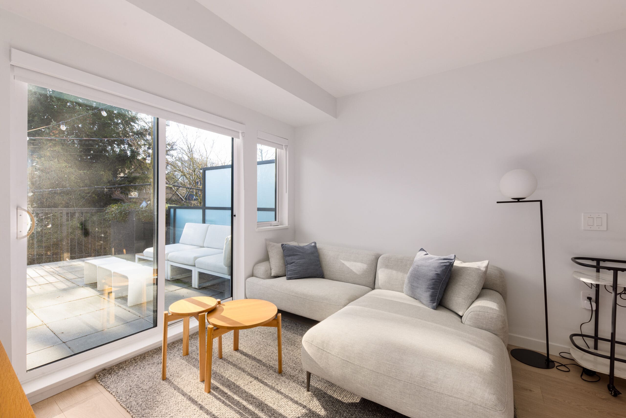 A bright living room with a light gray sectional sofa, small round wooden tables, a floor lamp, and sliding glass doors leading to a sunlit balcony with a white bench.