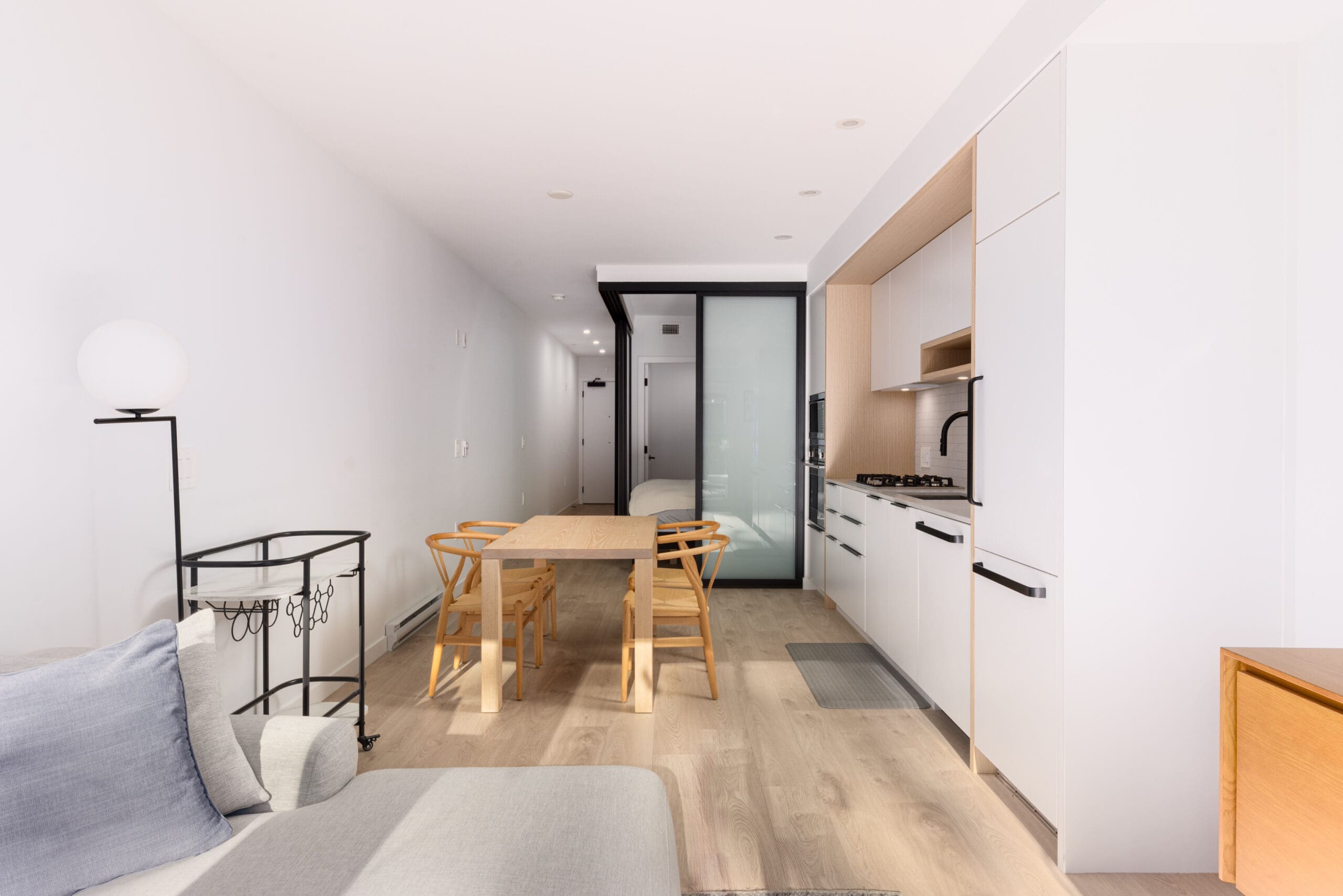 Minimalist studio apartment with light wood flooring, white walls, a kitchen area, small dining table with chairs, and a gray sofa. A black cabinet and modern light stand by the wall.