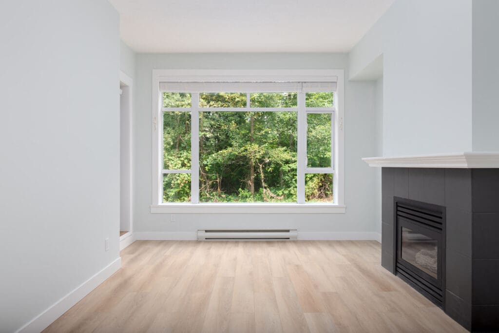 Empty room with pale wood flooring, large window with a view of greenery, and a dark-tiled fireplace on the right wall. Walls are painted light blue.