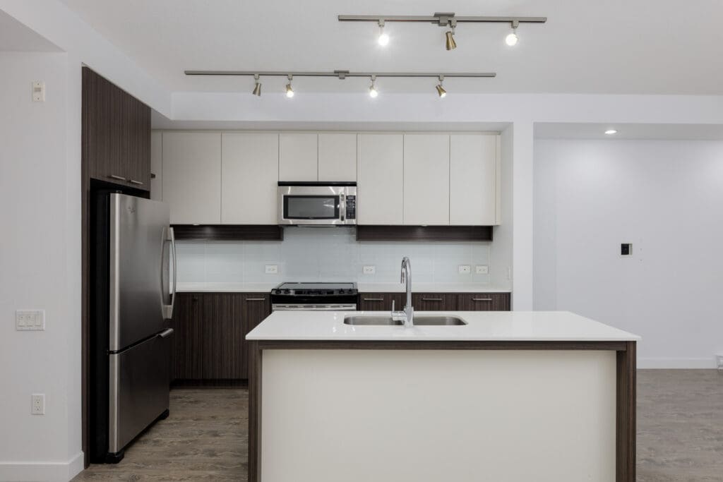 Modern kitchen with stainless steel appliances, white cabinets, and a center island featuring a sink. Track lighting is on the ceiling, and flooring is dark wood.