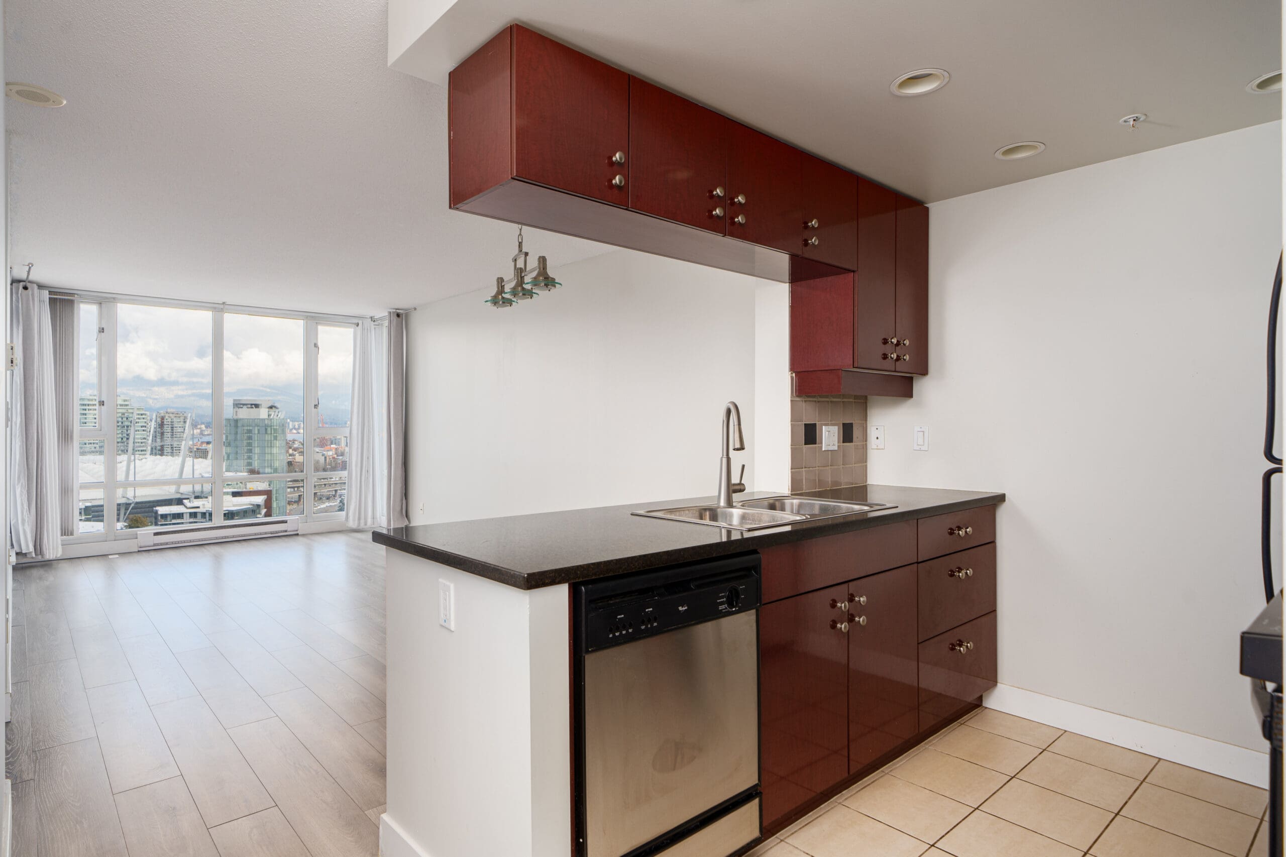 Modern open-plan kitchen with dark red cabinets, stainless steel appliances, and a sink. The space leads into a bright living area with large windows offering a city view.