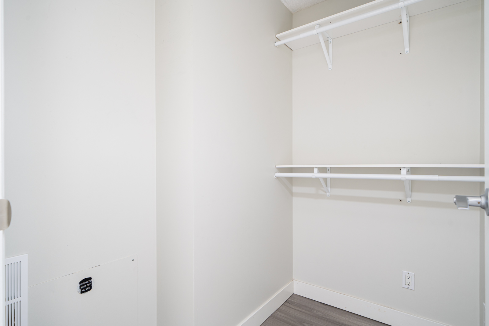 Empty white walk-in closet with wooden floor, two white hanging rods, and a small light switch on the right wall.