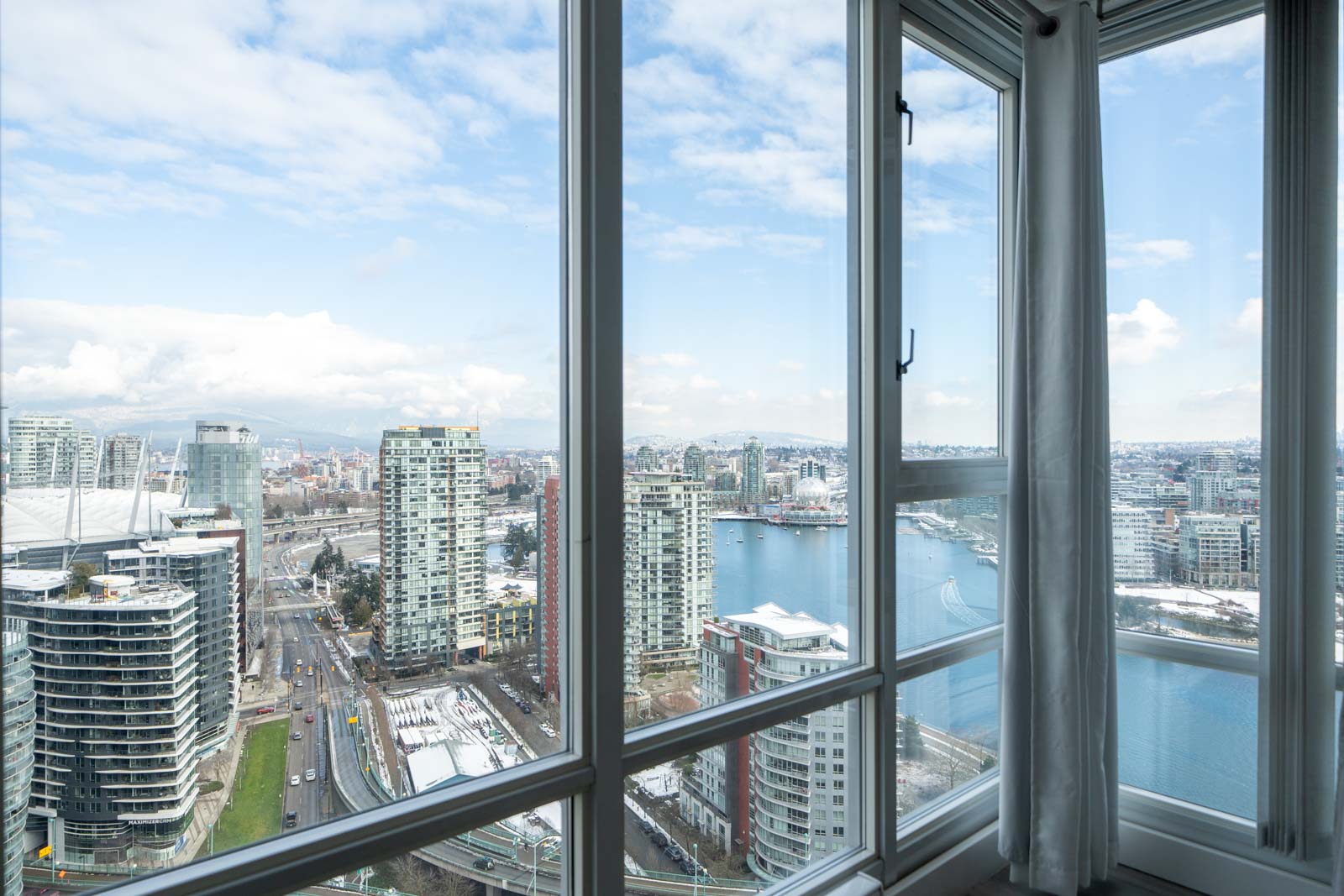 Cityscape view from a high-rise window showing buildings, roads, and a body of water under a cloudy sky.