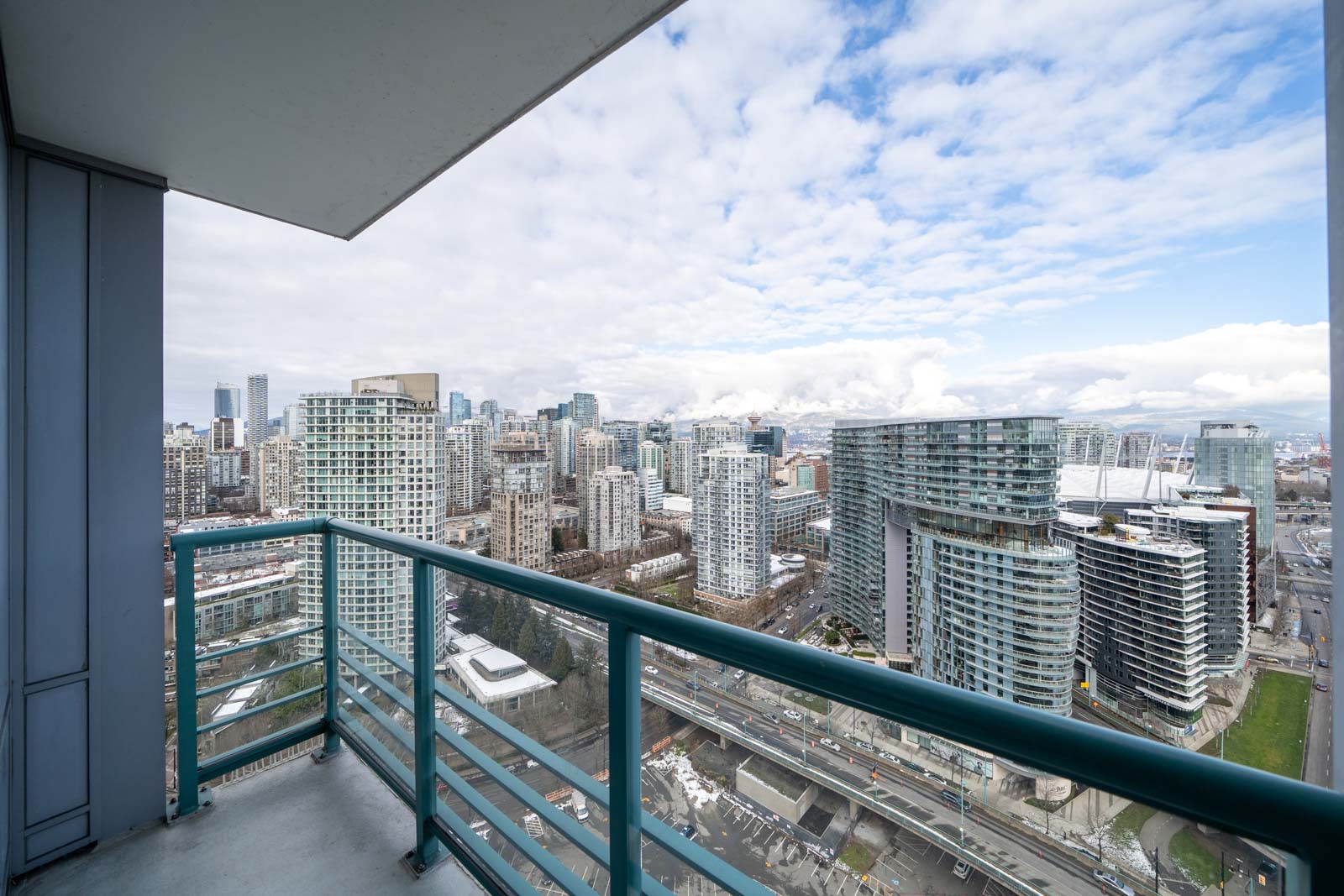 Urban skyline view from a high-rise balcony overlooking modern buildings, roads, and a partially cloudy sky.