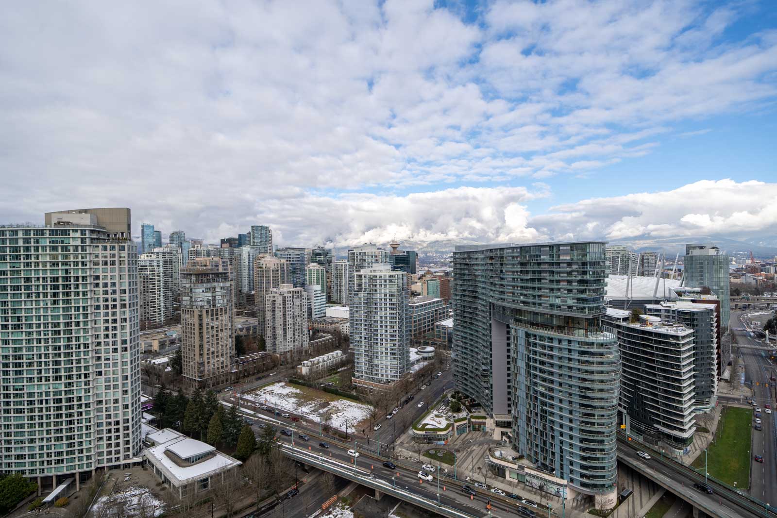 Aerial view of a cityscape with modern high-rise buildings, roads, and some snow patches. A partly cloudy sky is visible above.