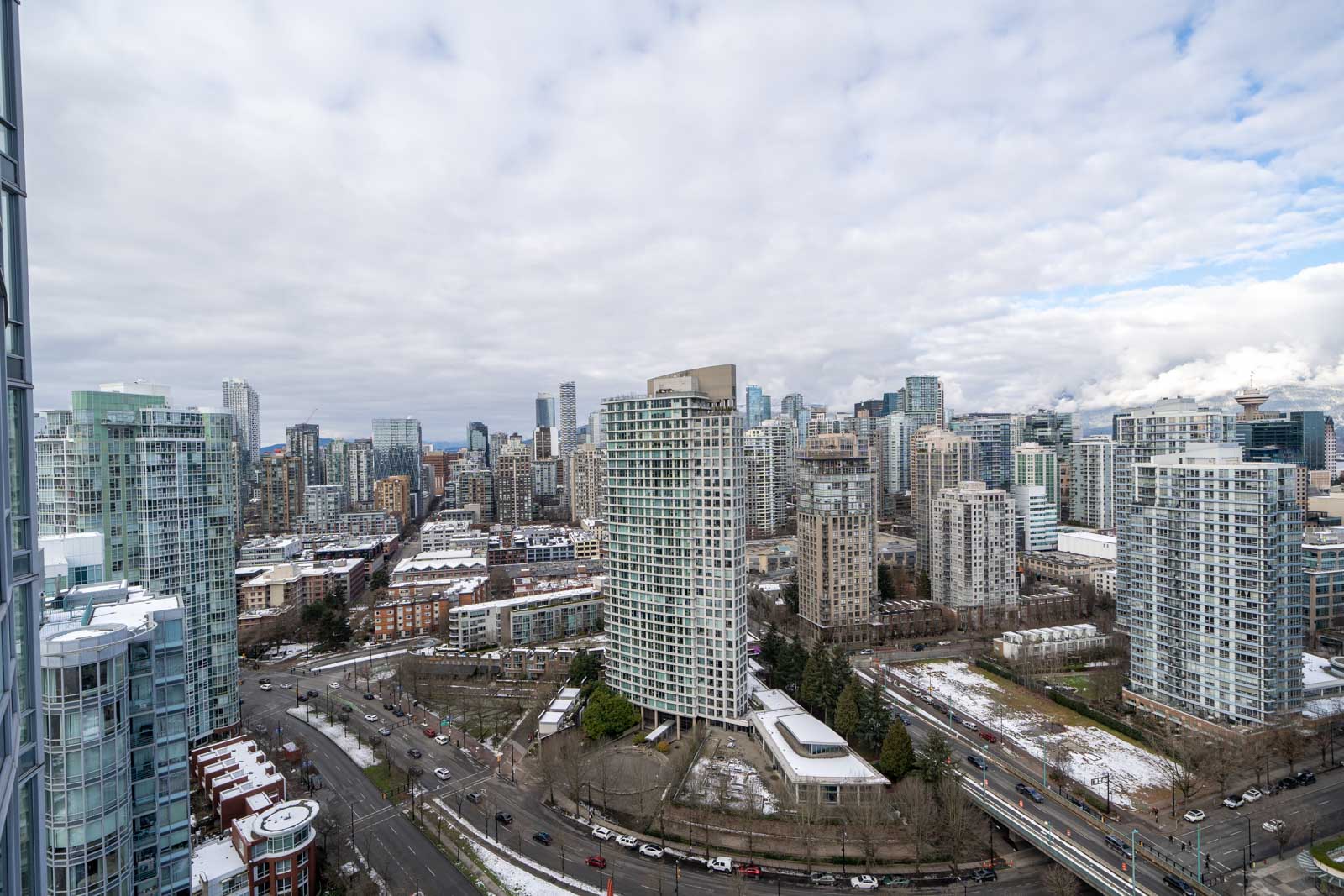 Aerial view of a cityscape with numerous high-rise buildings under a cloudy sky. Roads with light traffic curve through the area. Some snow is visible on rooftops and greenery.