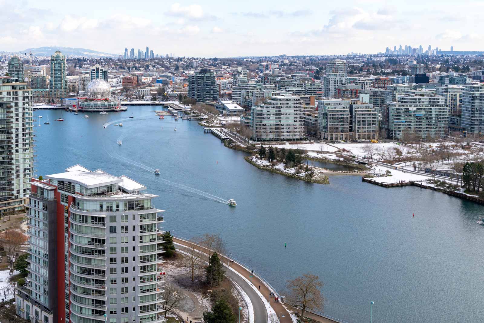 Aerial view of a cityscape with a river, surrounded by modern buildings, and light snow on the ground. Boats are visible on the water. Cloudy sky in the background.