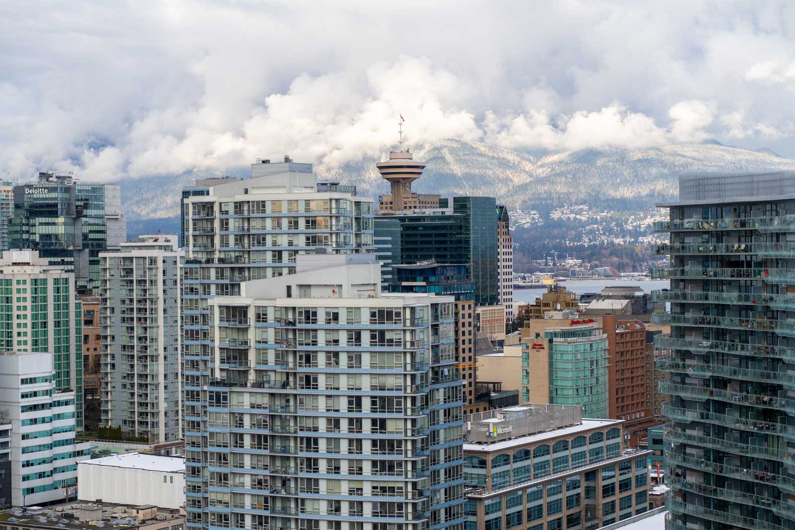 Cityscape view of a downtown area with modern high-rise buildings, a tower in the distance, and mountains partially covered with clouds in the background.