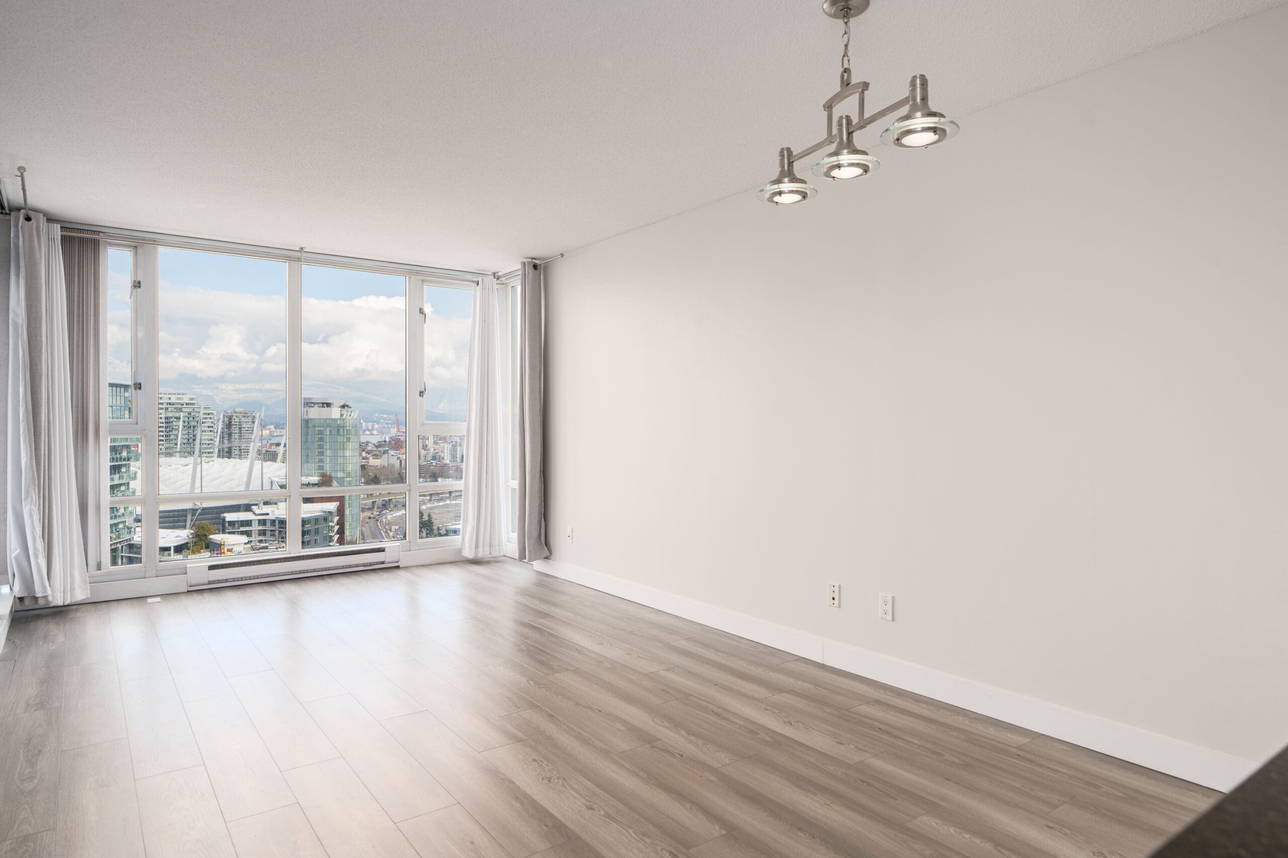 Bright, empty modern room with wooden flooring, large windows offering cityscape views, and a ceiling light fixture.