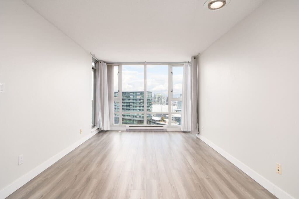 Empty room with light wood flooring, white walls, and large windows overlooking modern buildings and a cloudy sky.