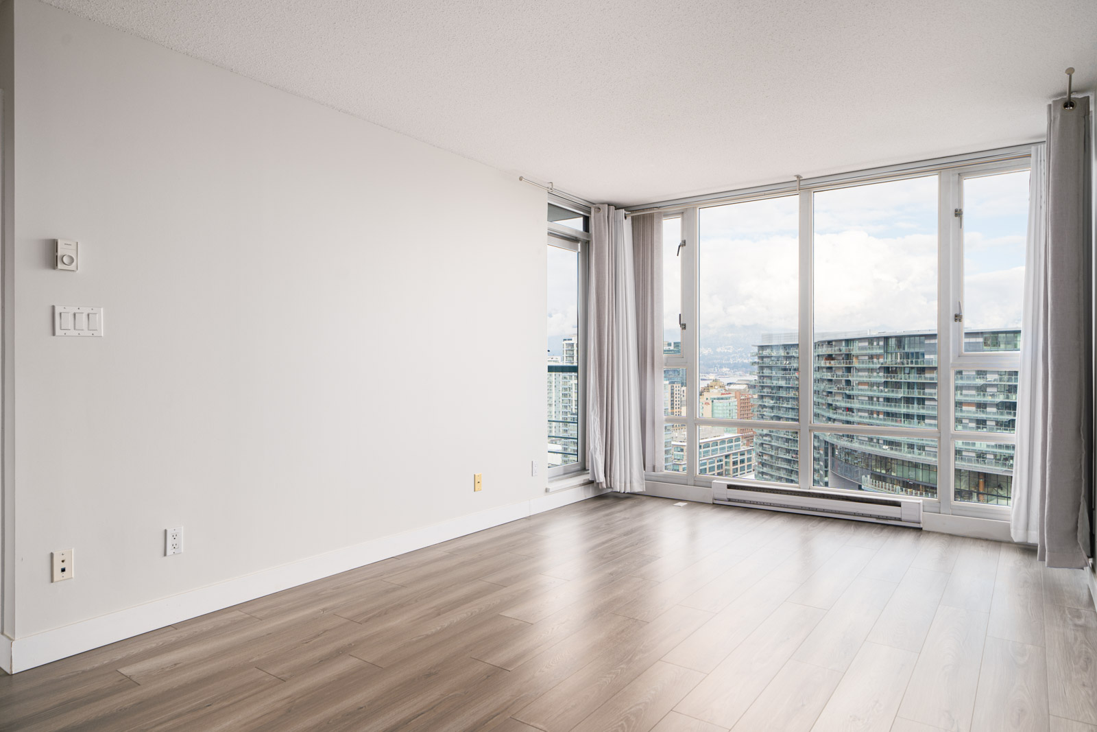 Empty modern apartment room with large windows, light wood flooring, white walls, and cityscape view.