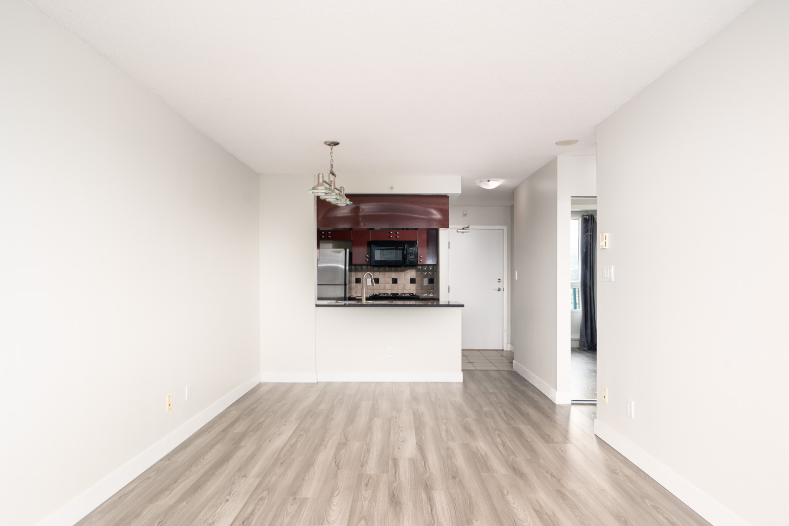 Empty living room with light wood flooring, white walls, and a view of a kitchen area featuring dark countertops, a microwave, and stainless steel appliances.