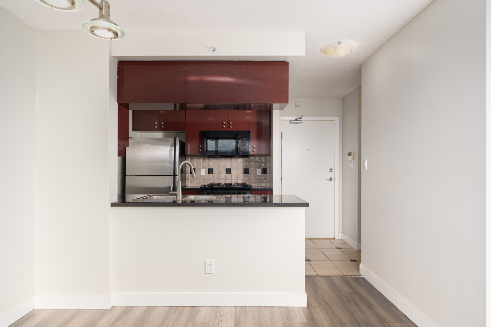 Modern kitchen with stainless steel appliances, dark wood cabinets, and a small island with a sink. Light wood flooring and a tiled entryway are visible.