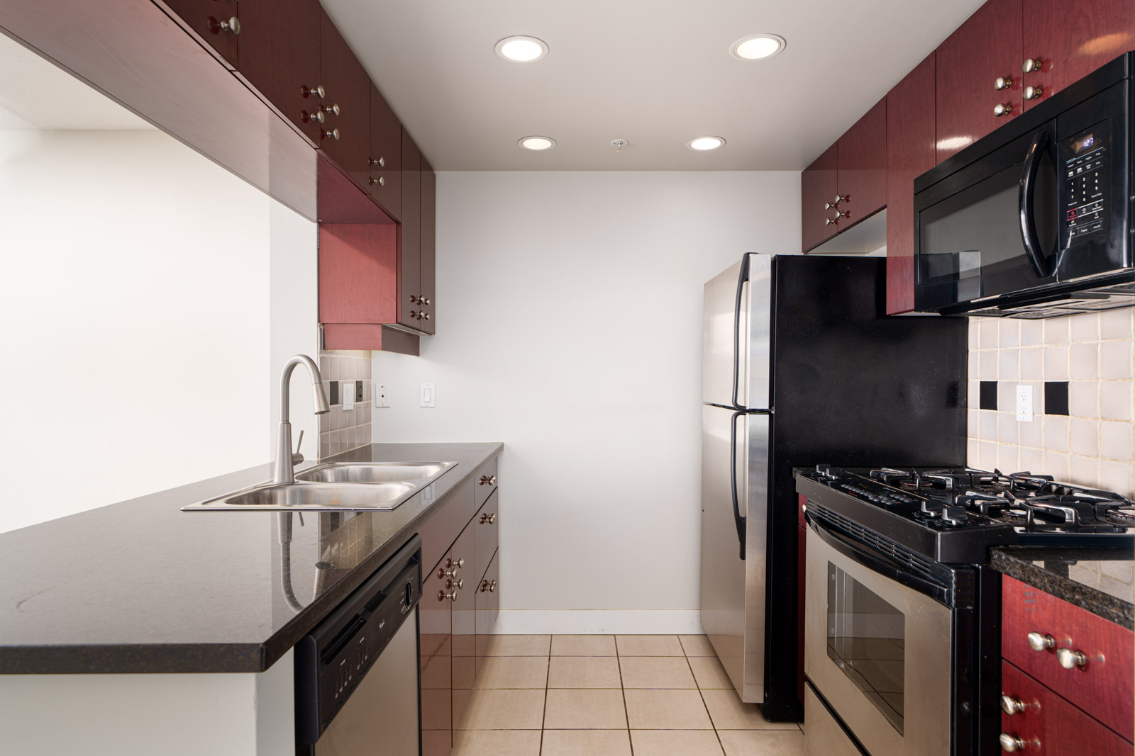 Modern kitchen with red cabinets, black countertops, stainless steel sink, dishwasher, refrigerator, gas stove, and microwave. Beige tile floor and white walls.