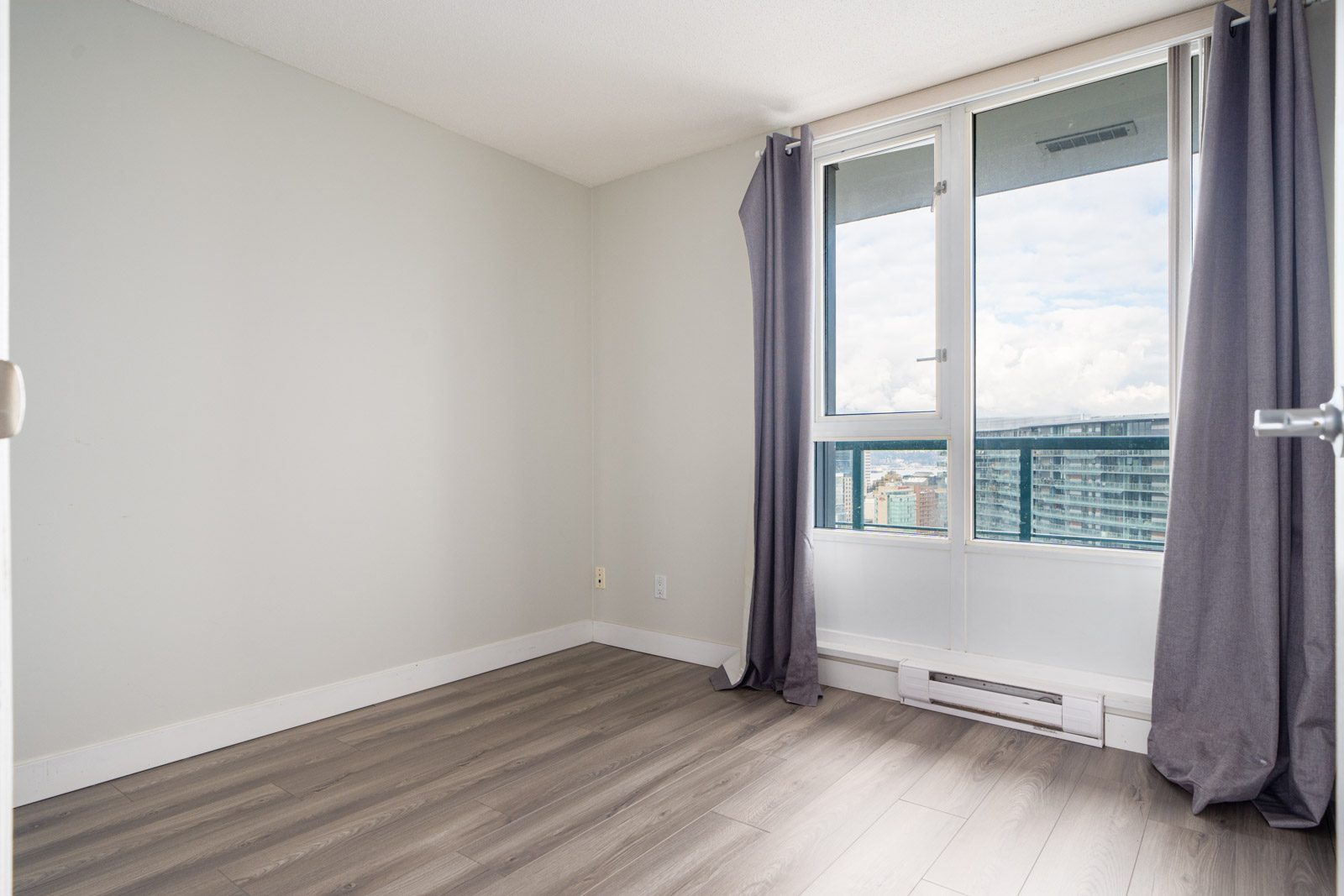 Empty room with light gray walls, hardwood floors, a large window with gray curtains, and a view of cityscape and sky.