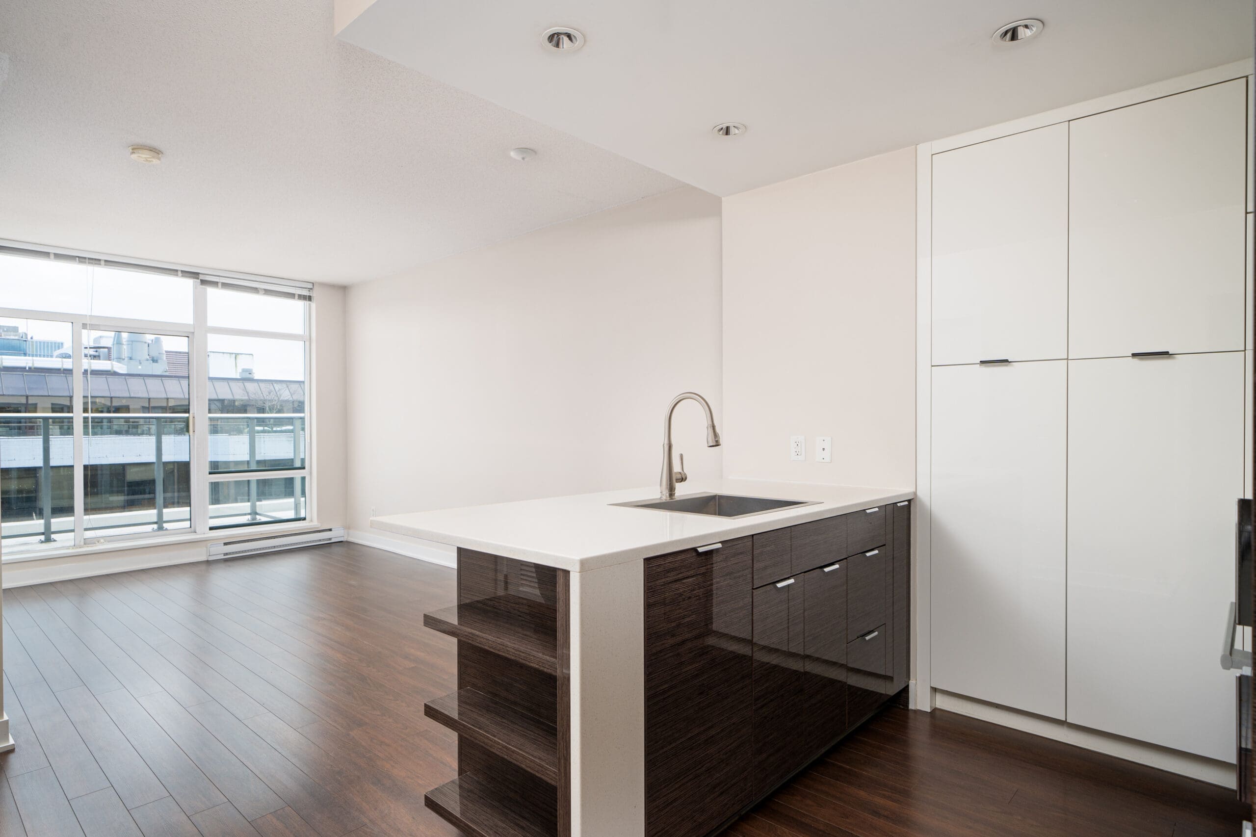 Modern apartment interior with a kitchen island featuring a sink and faucet, dark cabinetry, and wood flooring. Large window provides natural light.