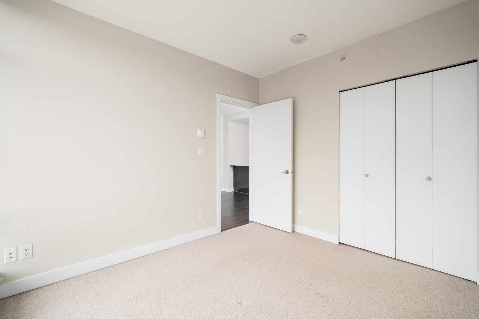 Empty room with beige carpet, white walls, and an open closet door. A single door leads to a hallway with dark flooring. Minimalist design with no furniture.