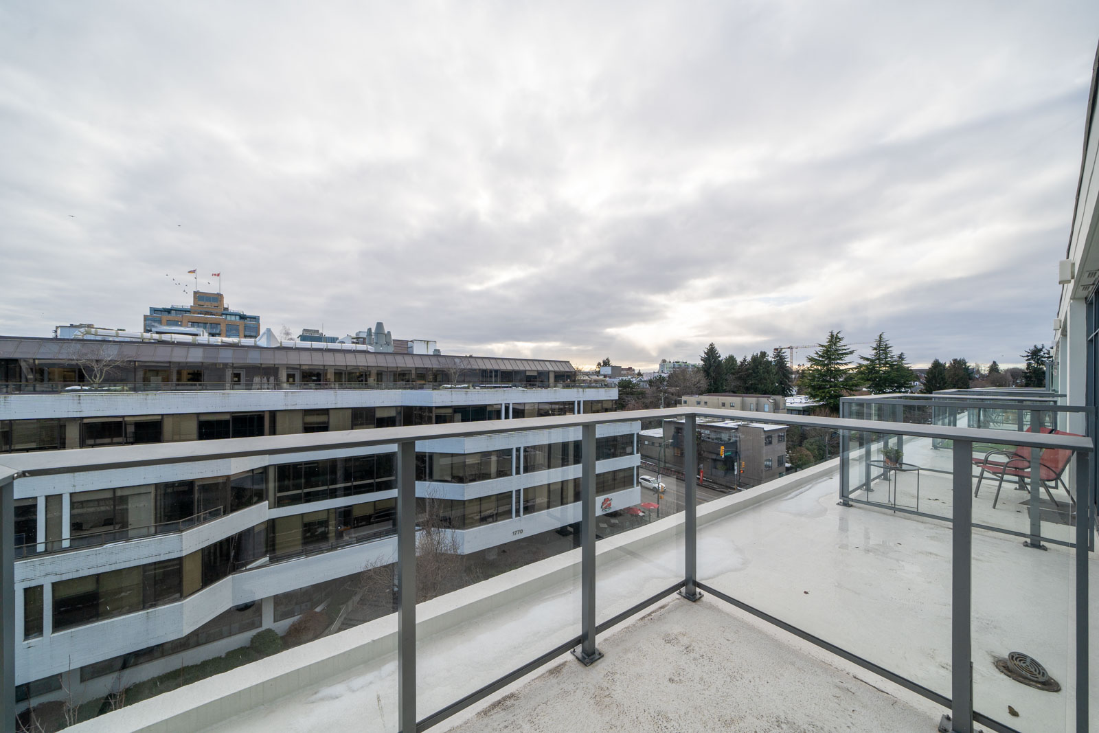 A rooftop view with glass railing, overlooking modern buildings under a cloudy sky.