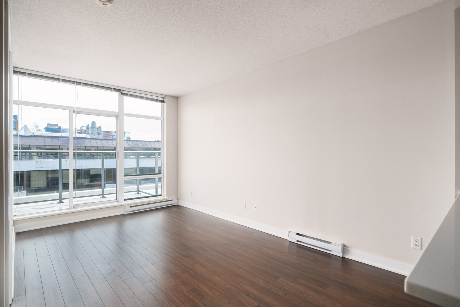 Empty room with large windows, hardwood floor, and white walls overlooking a cityscape.