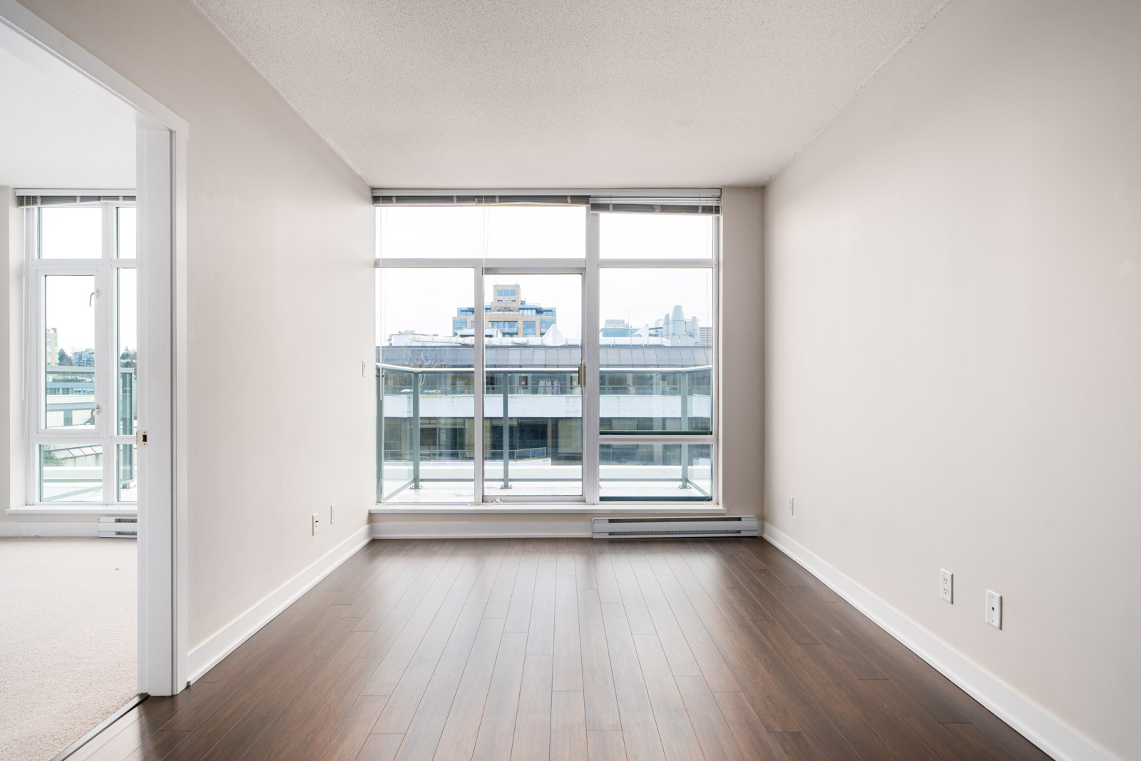 Empty room with large floor-to-ceiling windows, wooden floor, white walls, and a view of buildings outside. Doorway leads to an adjacent space.