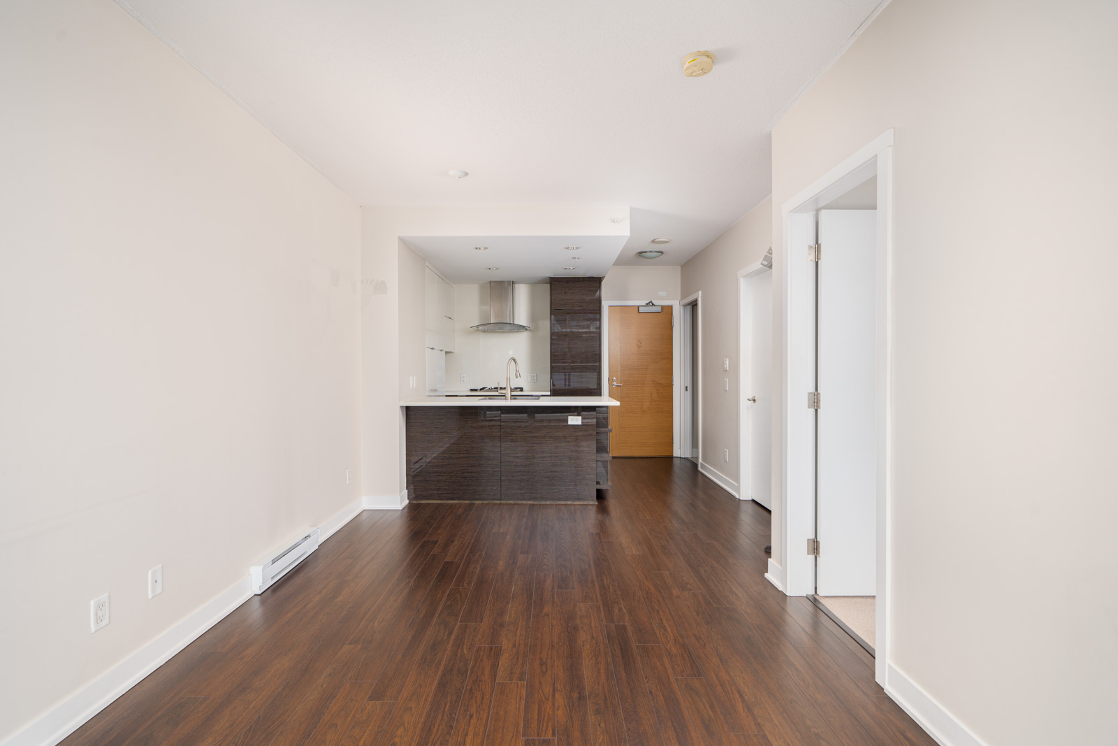 A modern, empty kitchen and living space with dark wood flooring, white walls, and a compact kitchen area featuring dark cabinets and stainless steel appliances.