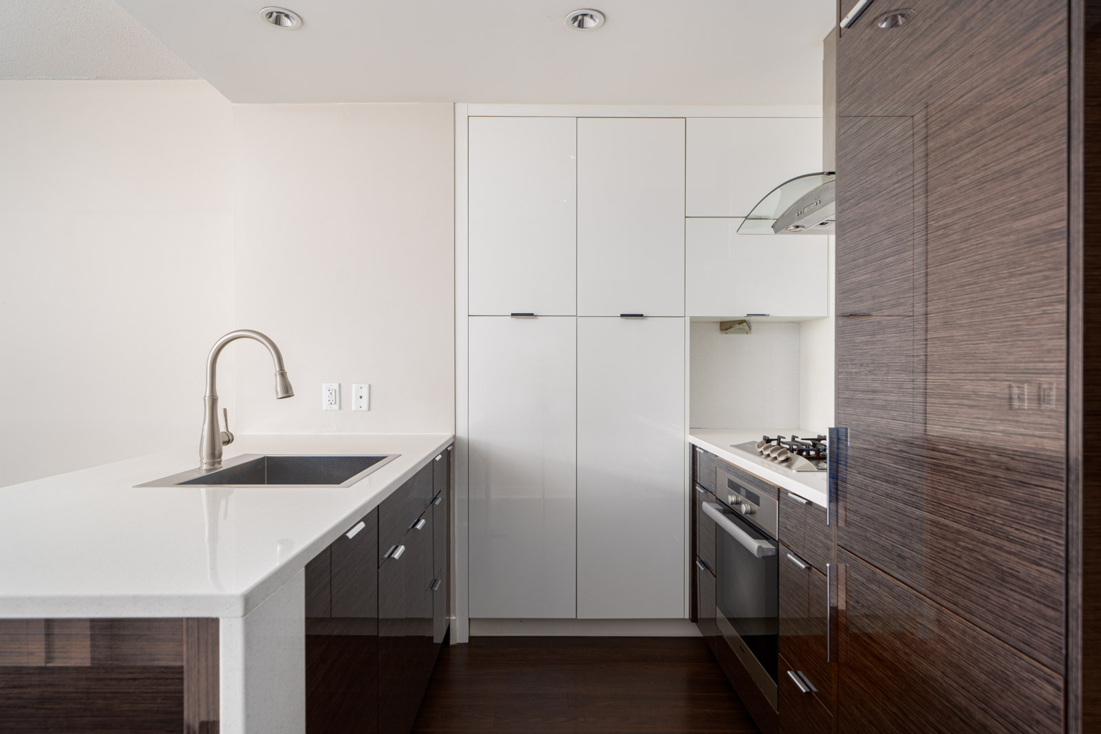 Modern kitchen with dark wood cabinets, white countertops, stainless steel faucet, and built-in appliances, including an oven and gas stove under a hood. Bright and minimalistic design.