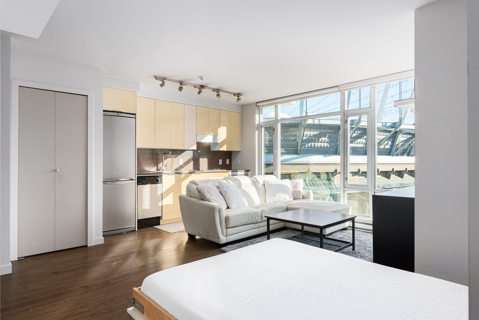 Bright, modern studio apartment with a small kitchen, white sofa, and large windows overlooking a stadium.