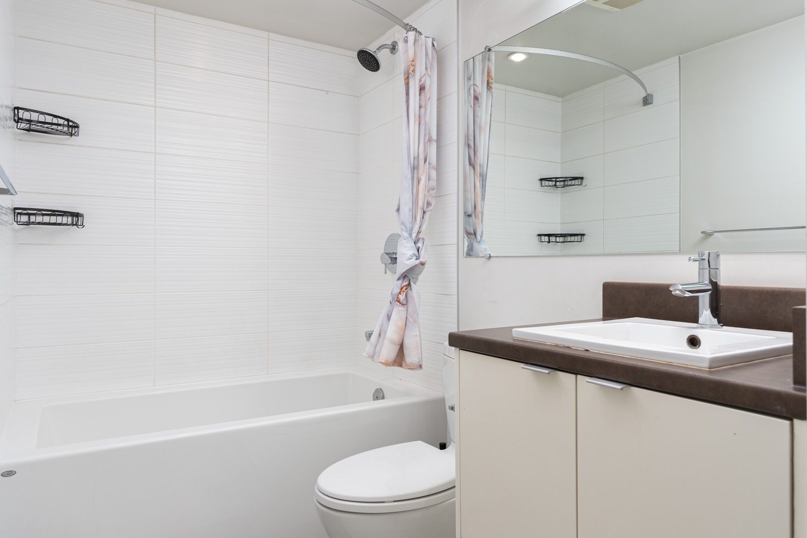Modern bathroom with a white bathtub, shower, toilet, and a sink with a mirror. The walls are tiled, and there is a shower curtain with patterns. Shelves are mounted on the walls.