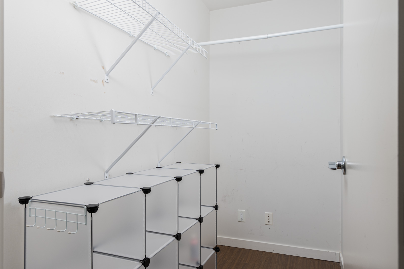 Empty closet with white wire shelves, a hanging rod, and white modular storage cubes. Wood floor and white walls with a closed door on the right.