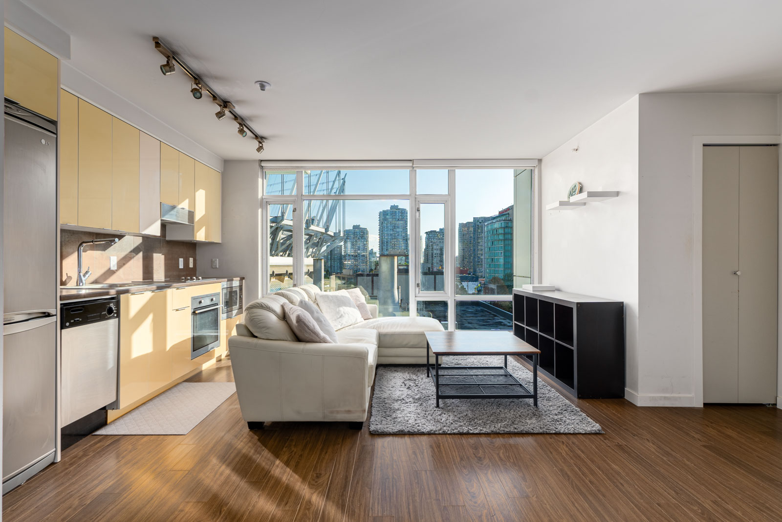 Modern living room with light wood flooring, a white sectional sofa, a black coffee table, and a wall of large windows overlooking a cityscape. Kitchen area with stainless steel appliances on the left.