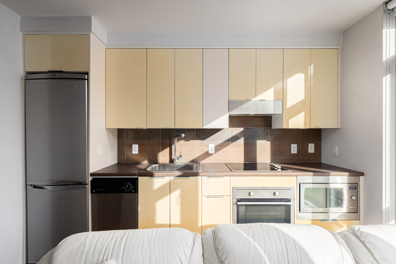 Modern kitchen with beige cabinets, black countertops, and stainless steel appliances, including a fridge, dishwasher, oven, and microwave. White couch in the foreground.