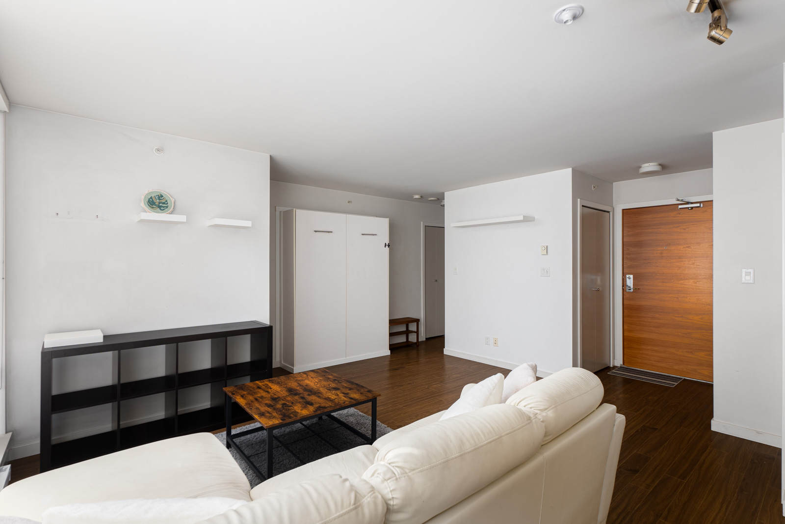 Minimalist living room with a white sofa, wooden coffee table, black shelving unit, and a brown door. White walls and hardwood floors create a clean, open space.