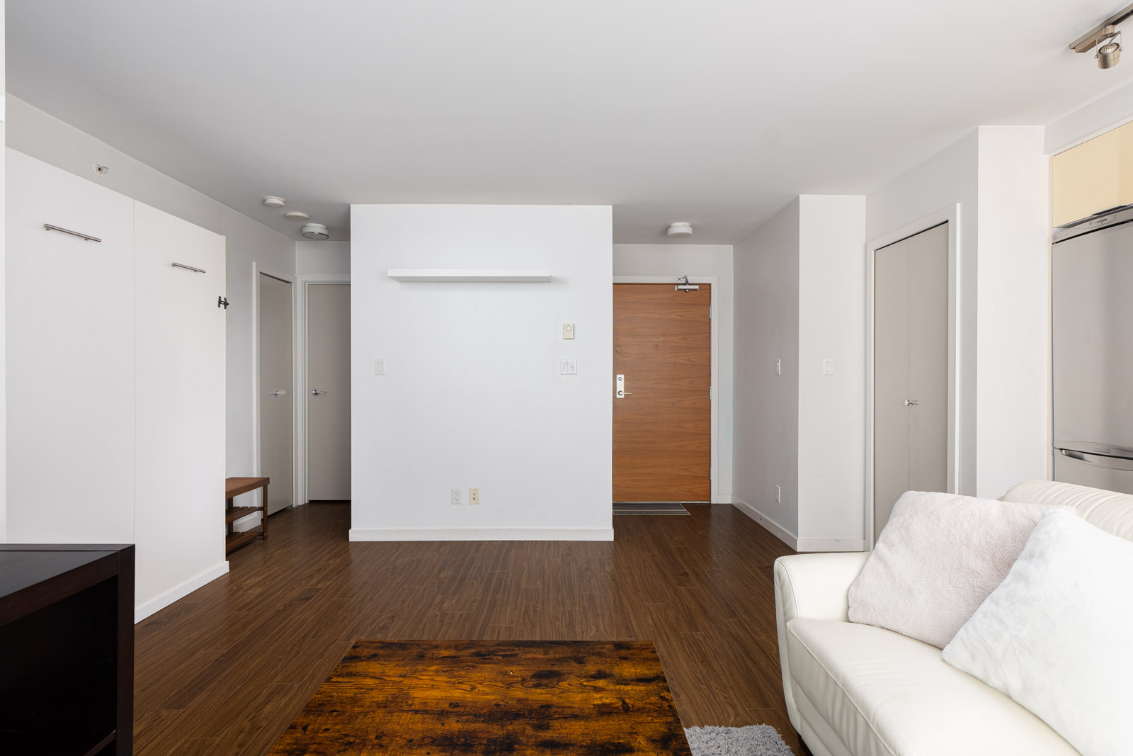 Minimalist living room with white walls, a white couch, wooden floors, and a wooden front door. A small rug lies on the floor, and the room features a few closed doors and cabinets.
