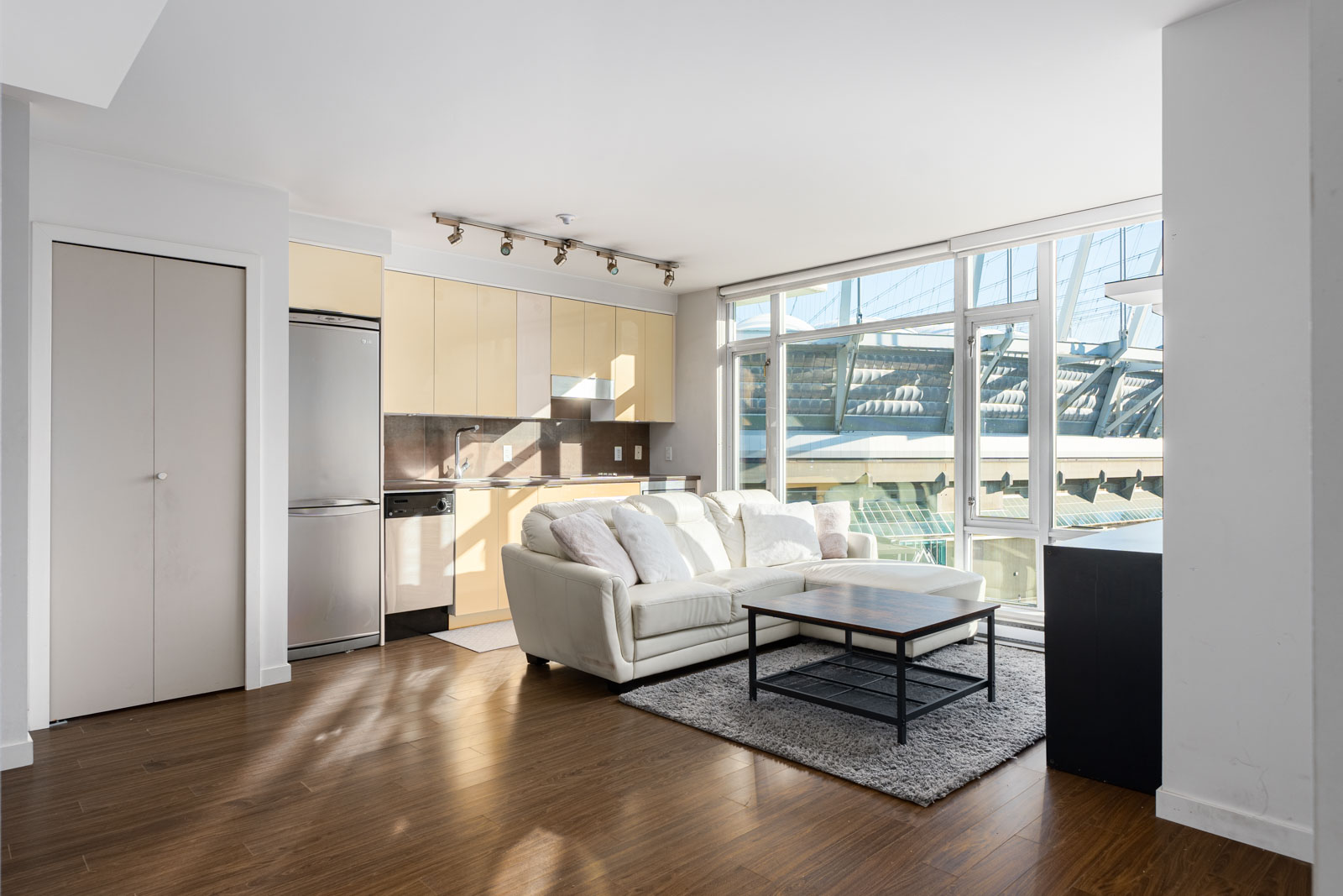 Modern apartment with large windows, a small kitchen, a white sofa, a black coffee table, and wooden flooring. Natural light fills the room.