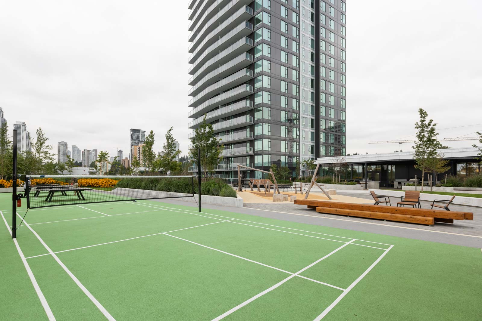 Rooftop with a tennis court, playground, and seating areas. A tall building is in the background with several other buildings visible in the distance.