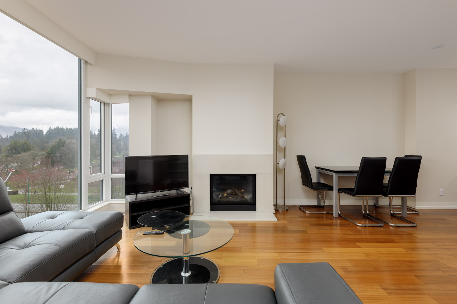Modern living room with hardwood floors, a sectional sofa, glass coffee table, TV, fireplace, and dining area with black chairs. Large window with a view of trees and cloudy sky.