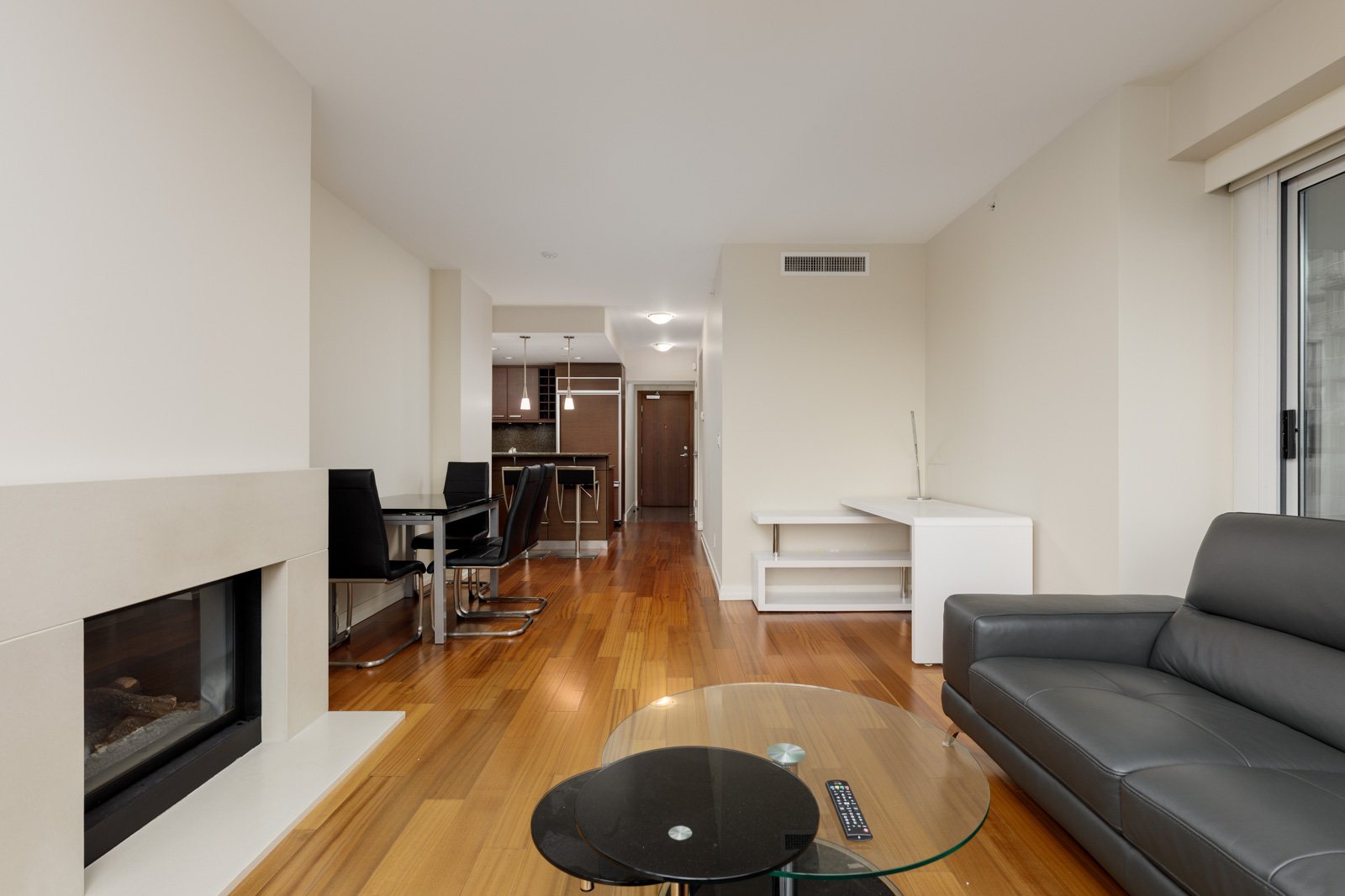 Modern apartment interior with a wooden floor, glass coffee table, gray sofa, dining area, fireplace, and open kitchen in the background.