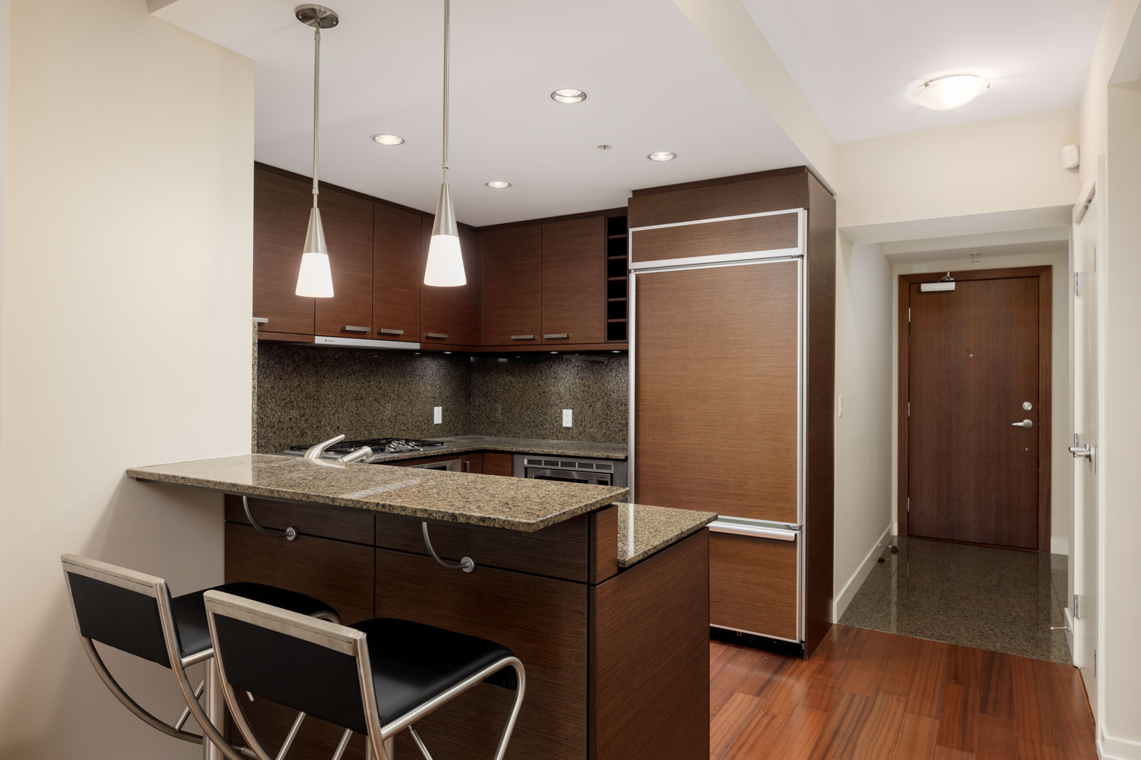 Modern kitchen with dark wood cabinets, granite countertops, and two pendant lights. Bar area with two chairs. Stainless steel appliances are visible. Wood flooring and a closed door in the background.