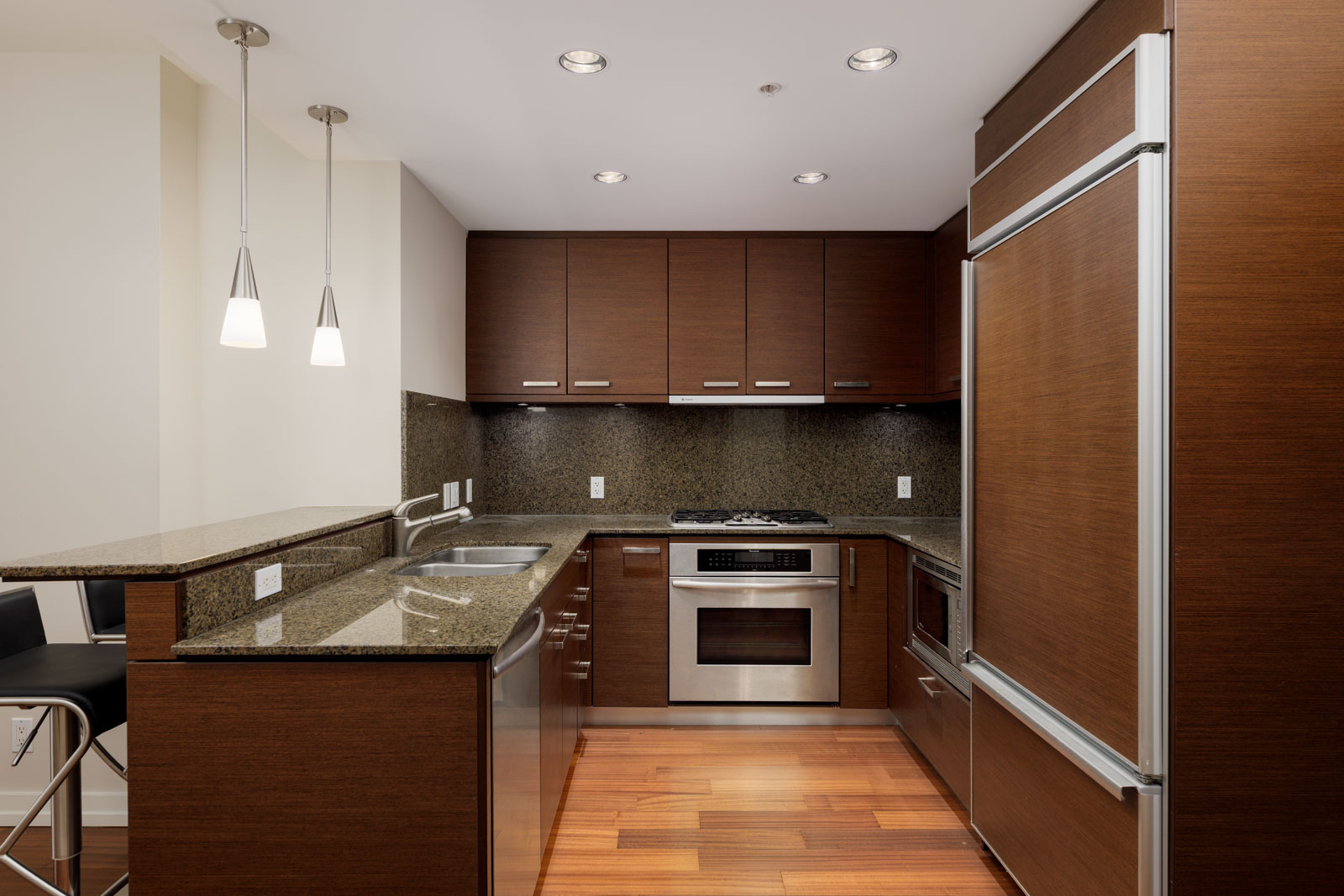 Modern kitchen with dark wood cabinets, granite countertops, and stainless steel appliances. Includes a built-in oven, stovetop, refrigerator, and pendant lights above a breakfast bar.