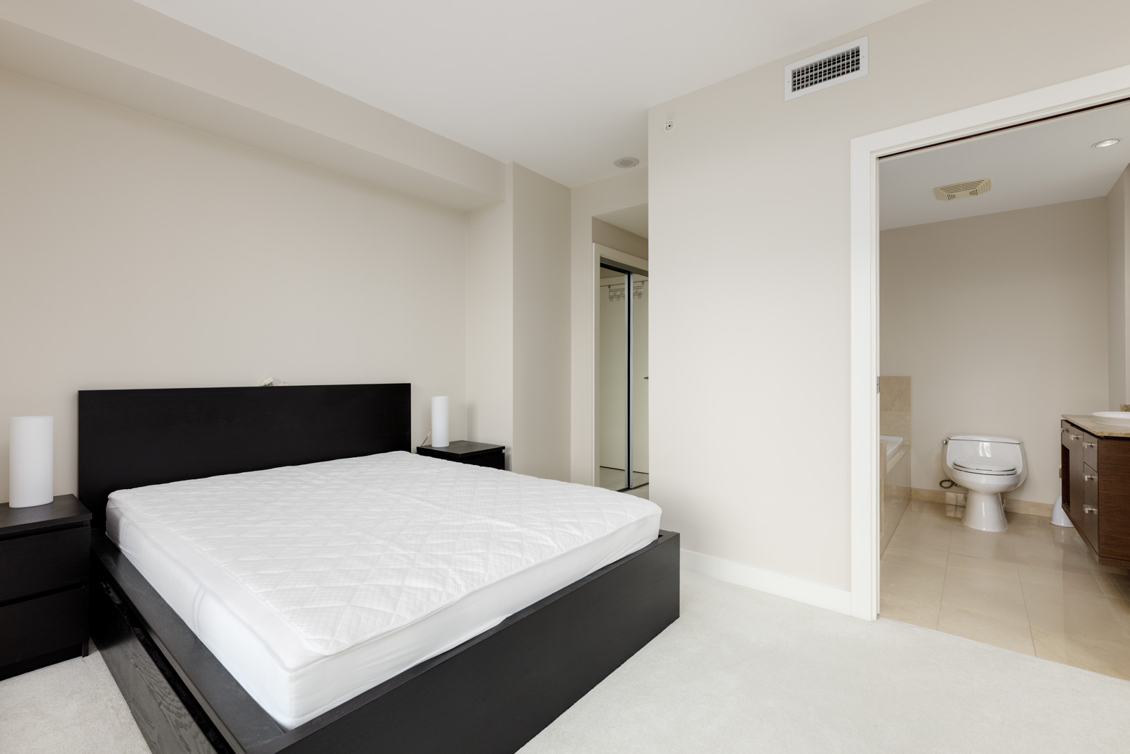 Bedroom with a black double bed frame and white mattress, two bedside tables, and a closet. An open door leads to a bathroom with a toilet and sink. Walls are beige, and flooring is light-colored.