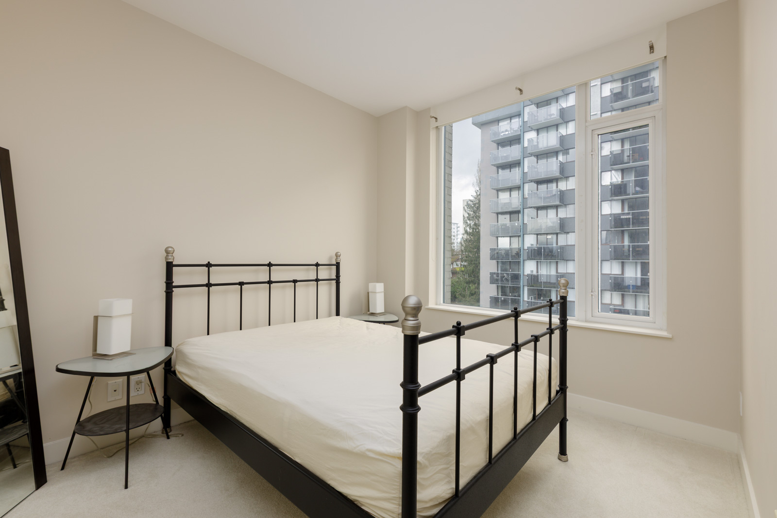 A bedroom with a metal frame bed, white bedding, a round side table with a lamp, and a large window showing a view of adjacent buildings.