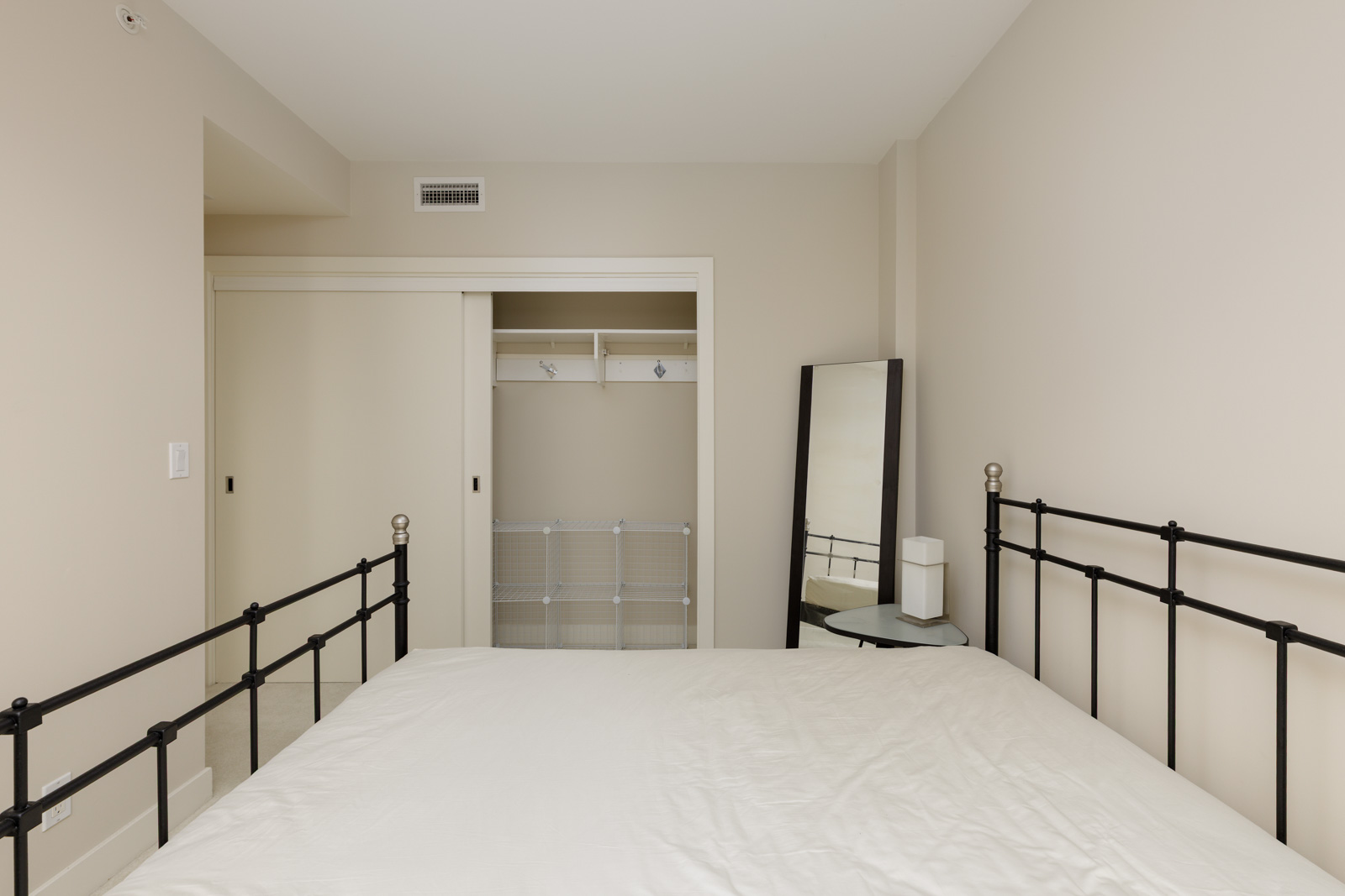 Minimalist bedroom with a metal bed frame, white bedding, open closet with sliding doors, standing mirror, and a small round table with a lamp. Walls are neutral-colored.