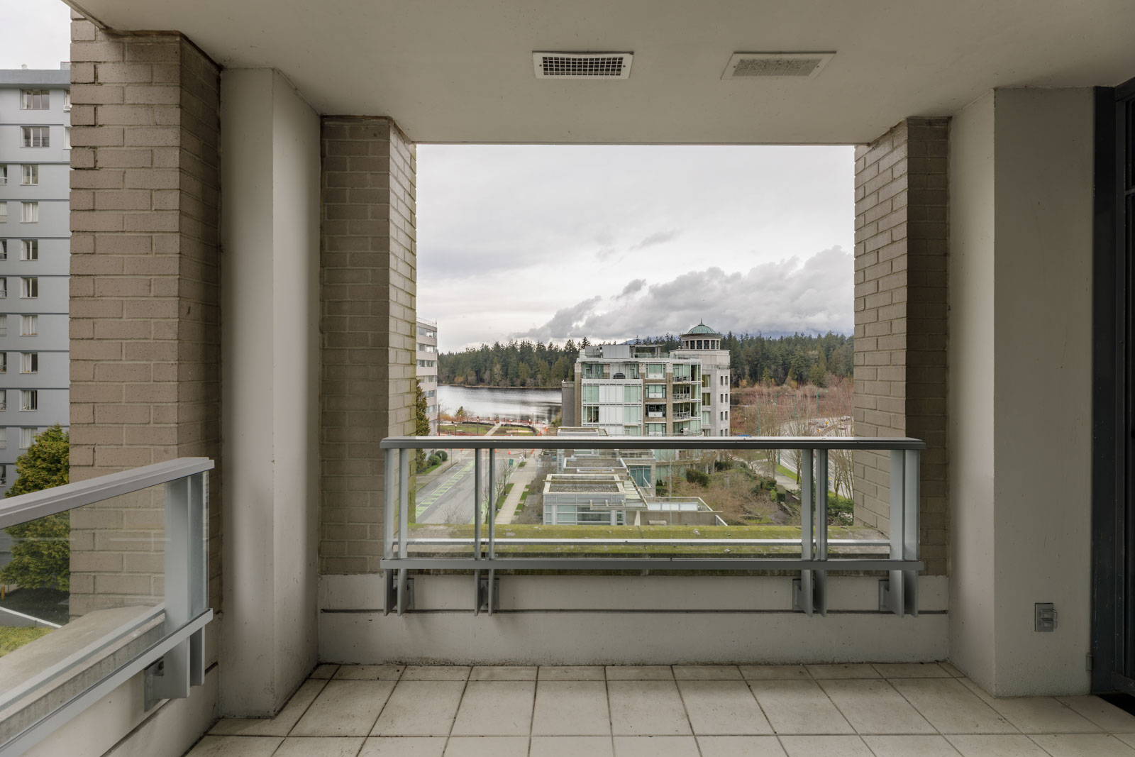 View from a balcony with a tiled floor, overlooking modern buildings, a road, and a distant body of water and trees under an overcast sky.