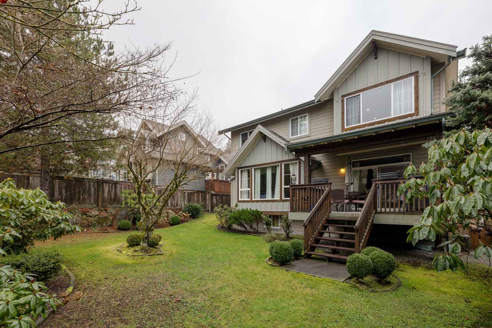 Two-story house with a wooden deck. Surrounded by a well-maintained yard, trees, and shrubs under a cloudy sky.