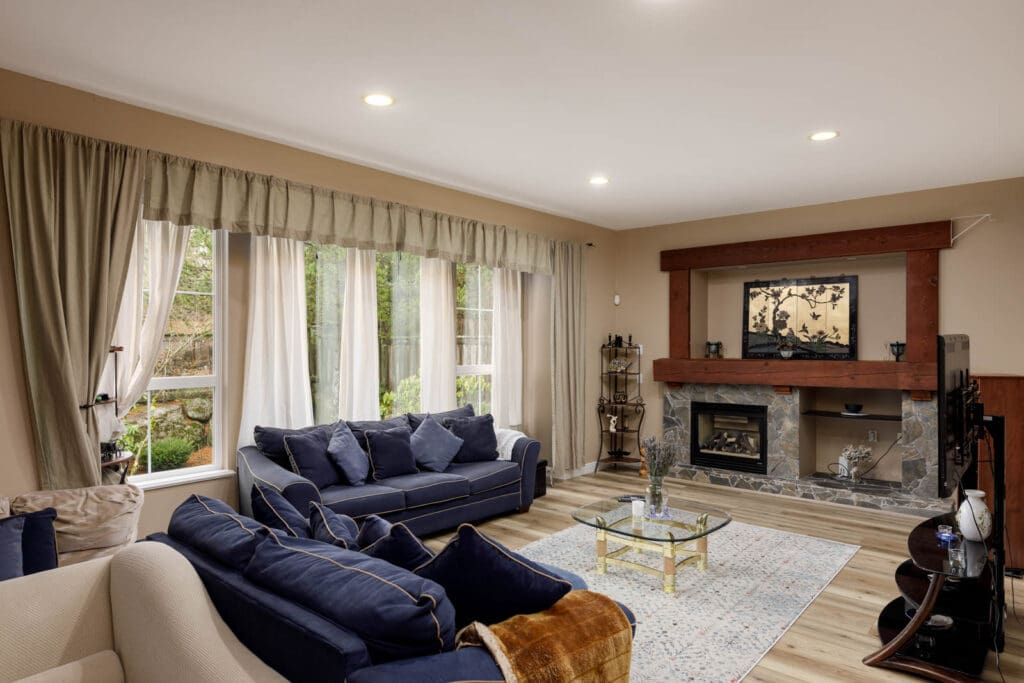 Living room with large windows, blue sofas, glass coffee table, fireplace, and a decorative wall piece above the mantel. Beige walls and curtains, wood floor, and recessed lighting.