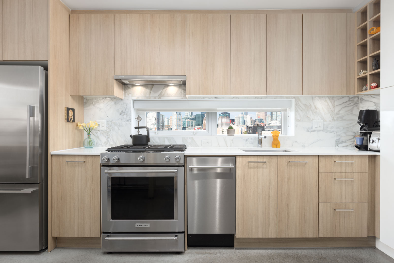 Modern kitchen with light wood cabinets, stainless steel appliances, and a marble backsplash. A large window above the counter offers a cityscape view. Flowers and decor items are visible.