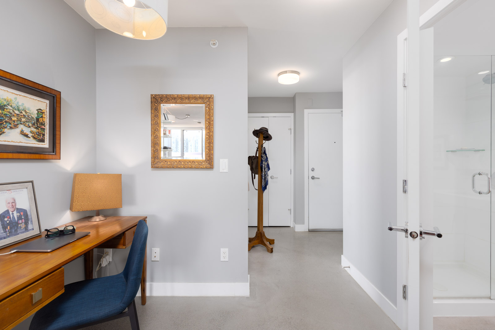 A small home office area with a wooden desk and blue chair, adjacent to a hallway and entry door. A lamp, framed picture, and mirror adorn the wall, with a standing coat rack nearby.